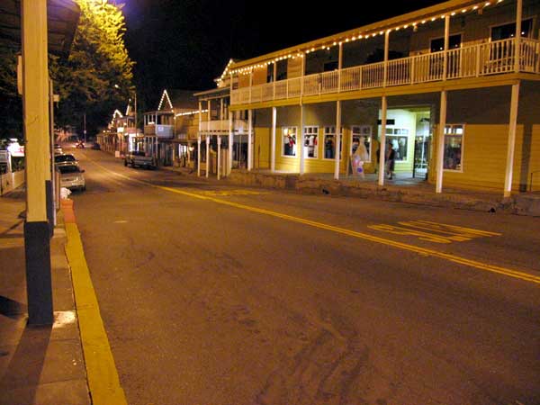 A quiet, empty street at night with lit storefronts and a two-story building with balconies evokes the timeless charm of Sutter Creek, where each corner tells a tale from the Days of Ore.