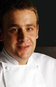 A smiling chef in a white jacket stands against a dark background, representing Tubal Restaurant in Tafalla, where Chef Nicolás Jimenez showcases his culinary expertise.